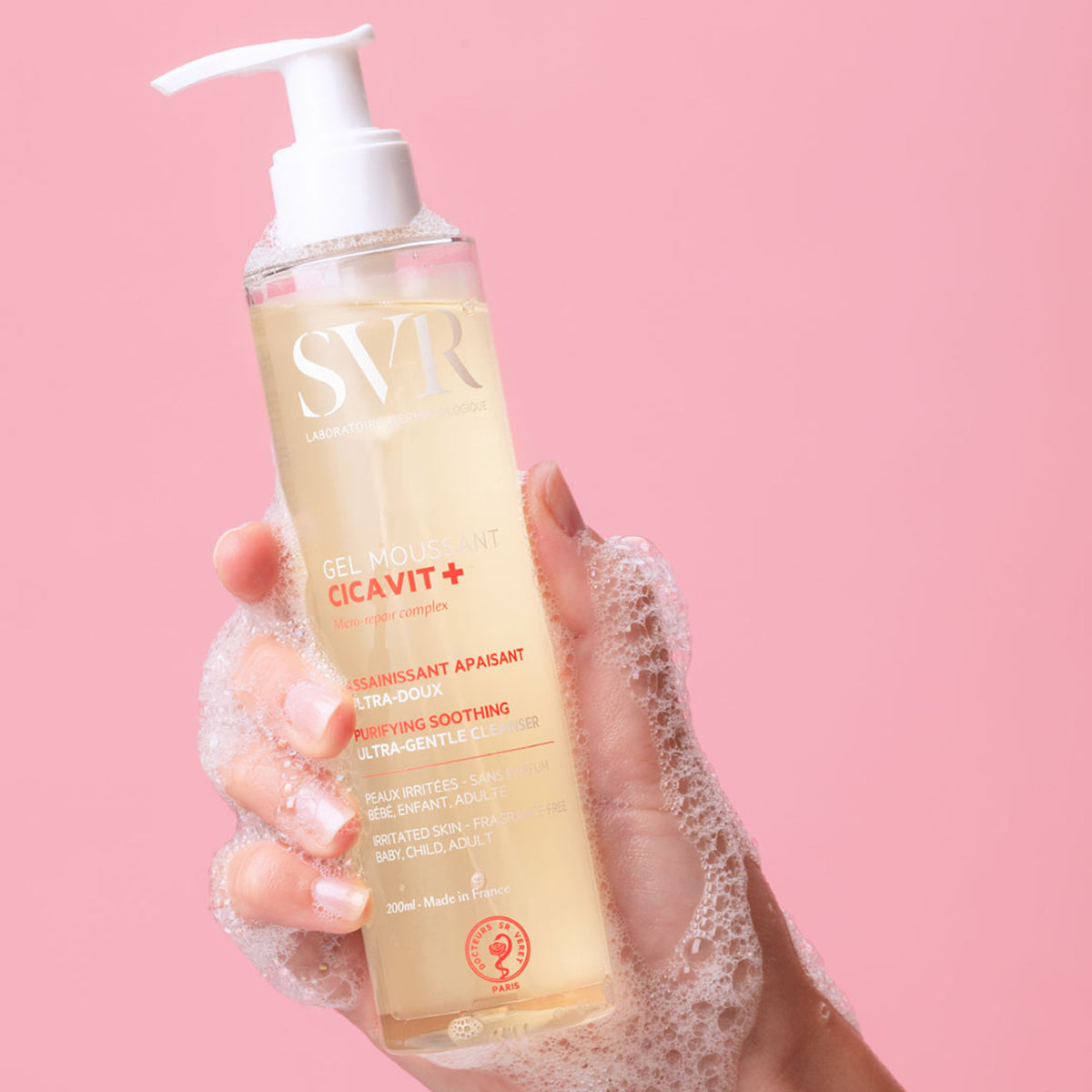 Hand hält eine transparente 200ml SVR Cicavit+ Gel Moussant Flasche mit weißem Pumpspender vor rosa Hintergrund.