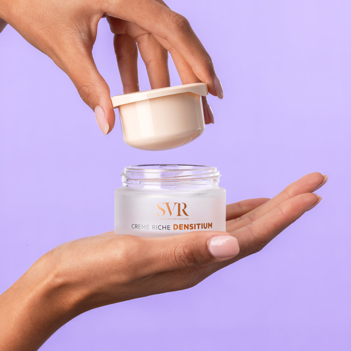 Hands holding an open jar of SVR DENSITIUM Crème riche with a frosted glass base and a pale beige lid against a purple background.