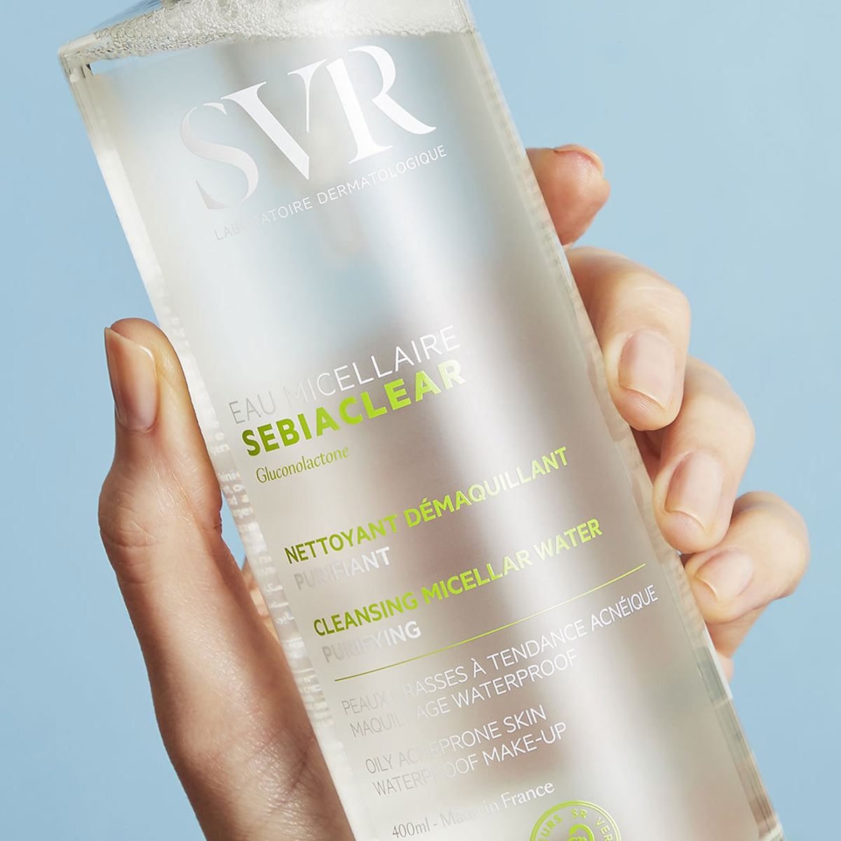 Transparente 400ml SVR SEBIACLEAR Mizellenwasser-Flasche mit weißer und grüner Schrift in einer Hand vor blauem Hintergrund.