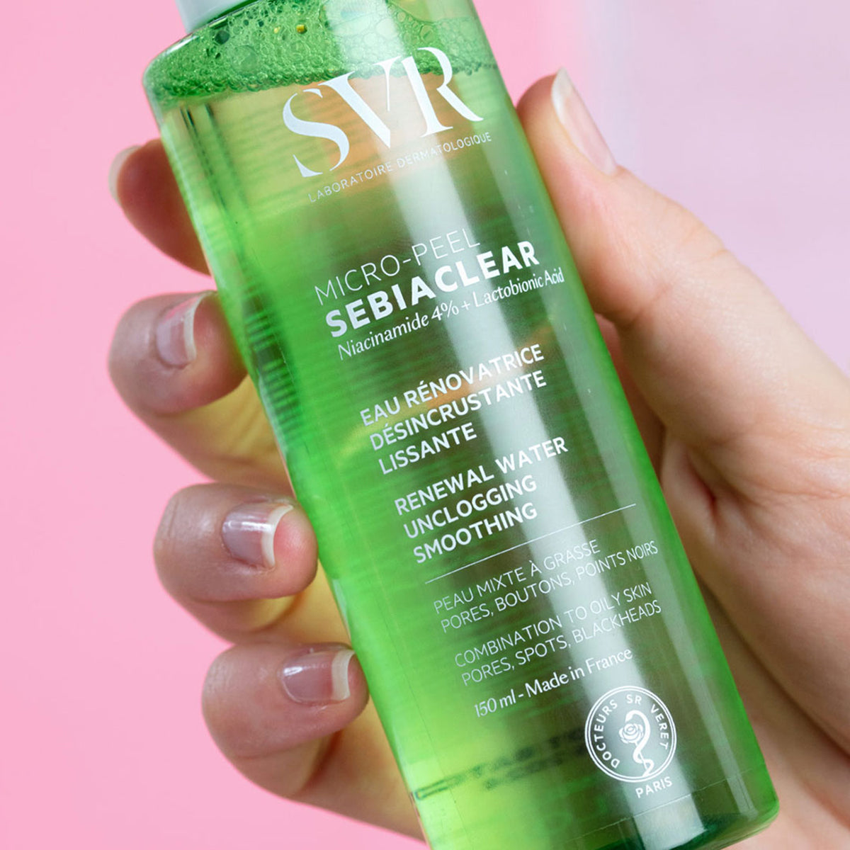 Transparente grüne SVR Sebiaclear Micro-Peel Wasserlotion in einer Hand vor einem rosa Hintergrund.