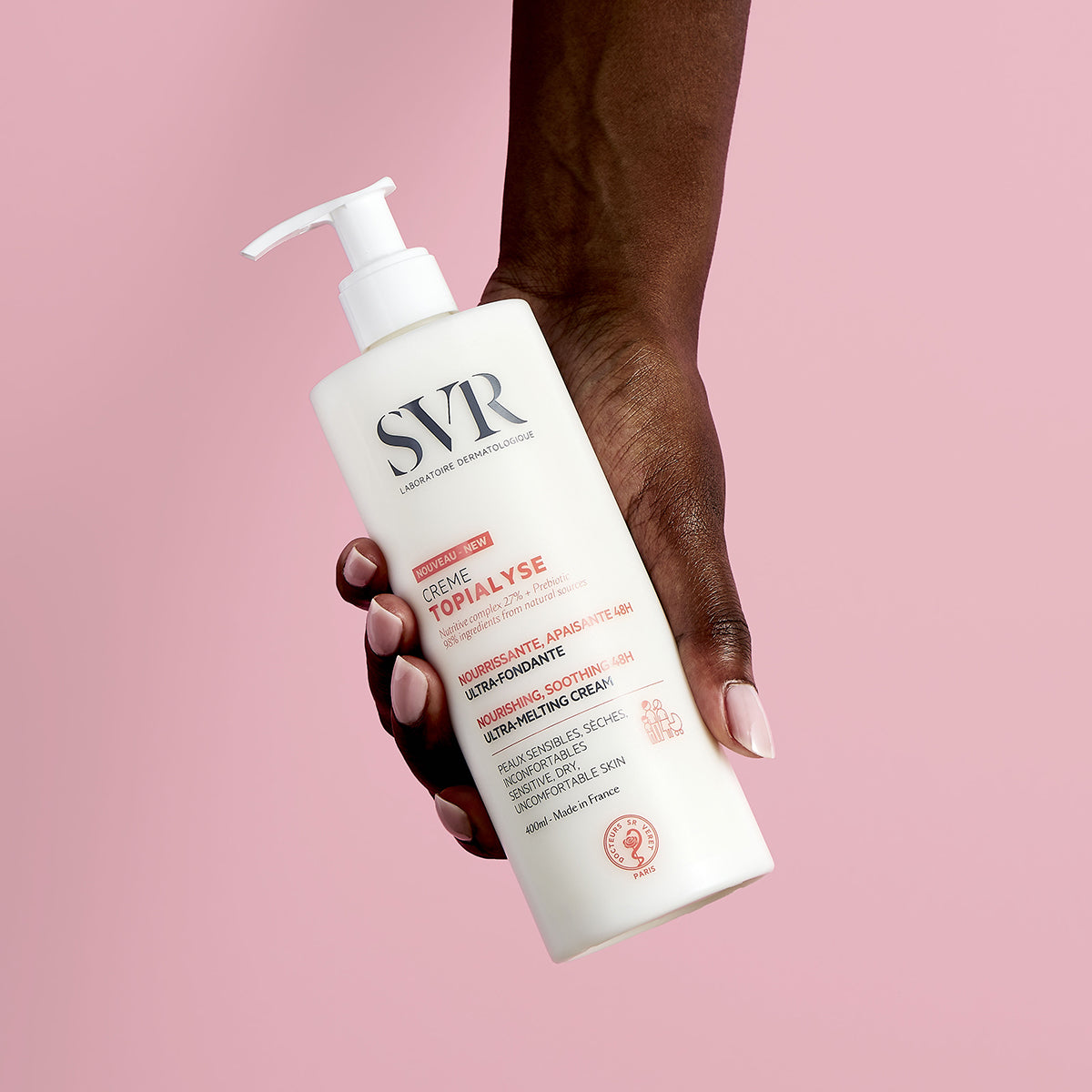 Hand hält weiße 400ml SVR TOPIALYSE Creme Pumpflasche vor einem hellrosa Hintergrund.