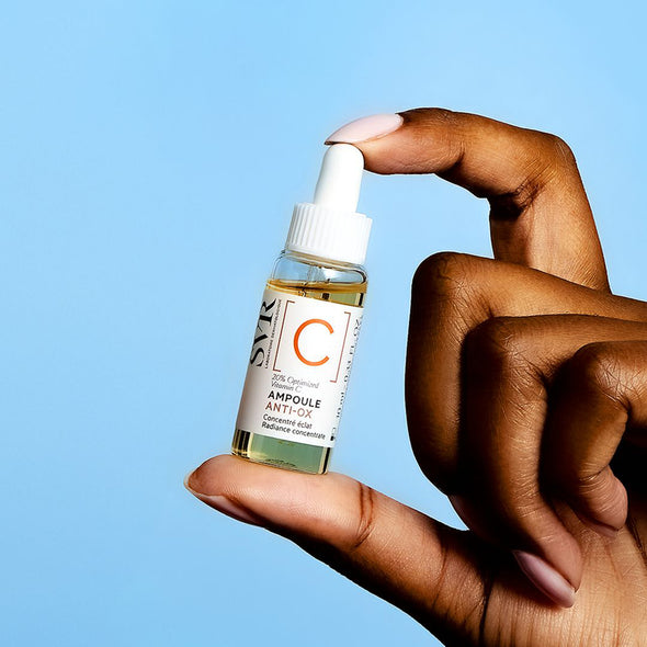 Hand hält eine kleine Glasflasche SVR Ampoule Anti-Ox Vitamin C Serum mit weißem Pipettenverschluss vor blauem Hintergrund.
