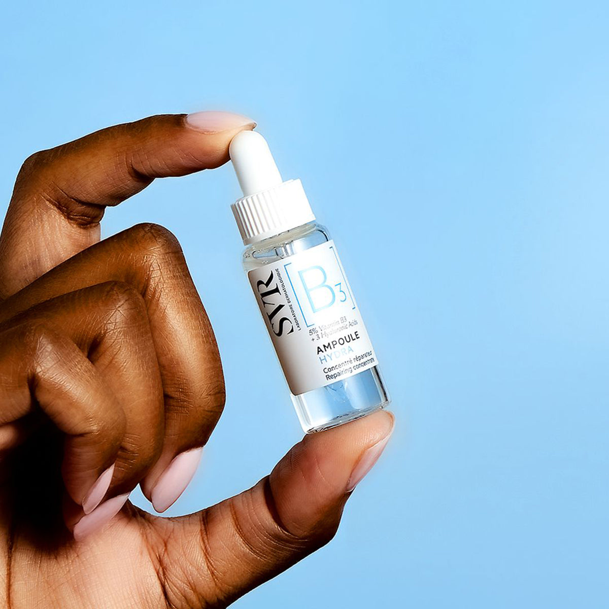 Hand hält eine kleine, transparente SVR B3 Ampoule Hydra Glasflasche mit weißem Pipettenverschluss vor blauem Hintergrund.