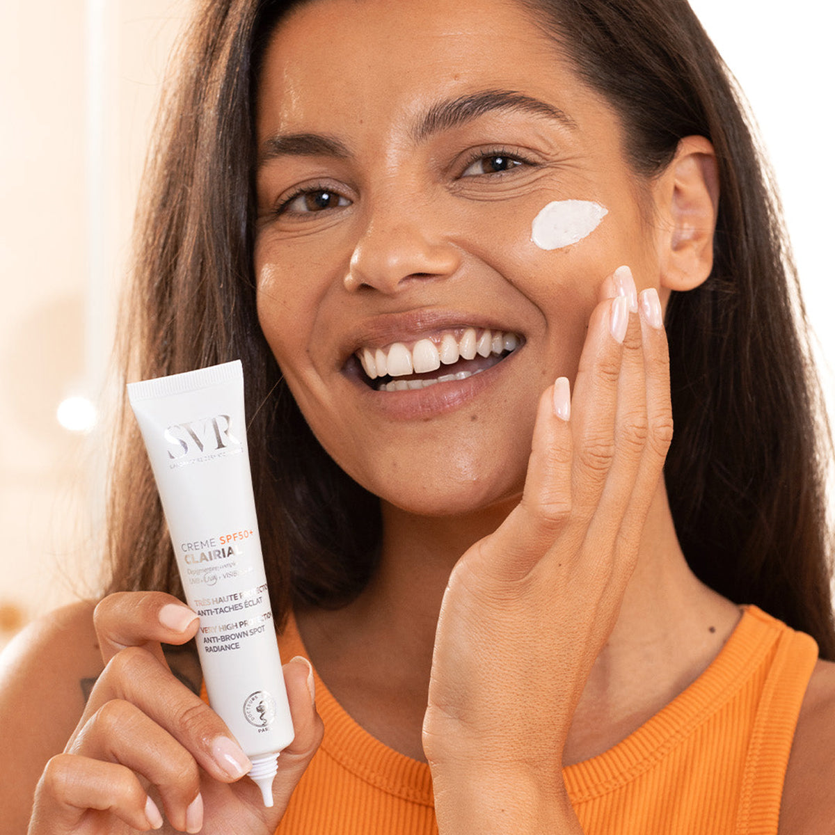 Lächelnde Frau hält eine weiße SVR Clairial Crème SPF50+ Tube und trägt die Creme auf ihre Wange auf.