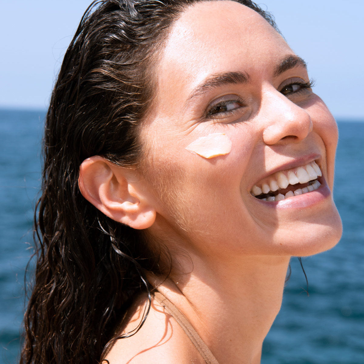 Lächelnde Frau mit nassen Haaren und einem Klecks aprikosenfarbener Sun Secure Blur SPF50+ Sonnencreme auf der Wange am Meer.