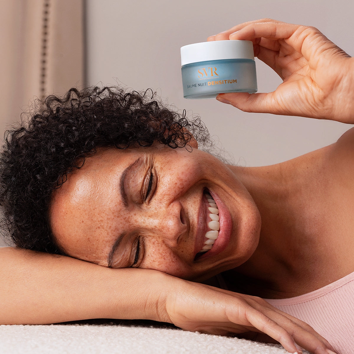 Smiling woman holding a frosted blue 50ml SVR Densitium Baume Nuit jar with a white lid against a soft background.
