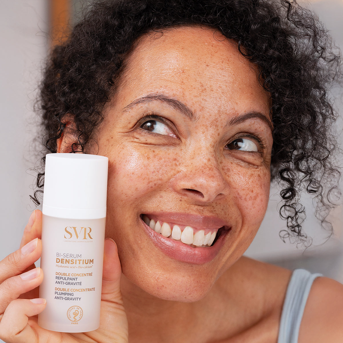 Smiling woman holding a white SVR Densitium Bi-Sérum bottle with gold text against a blurred background.