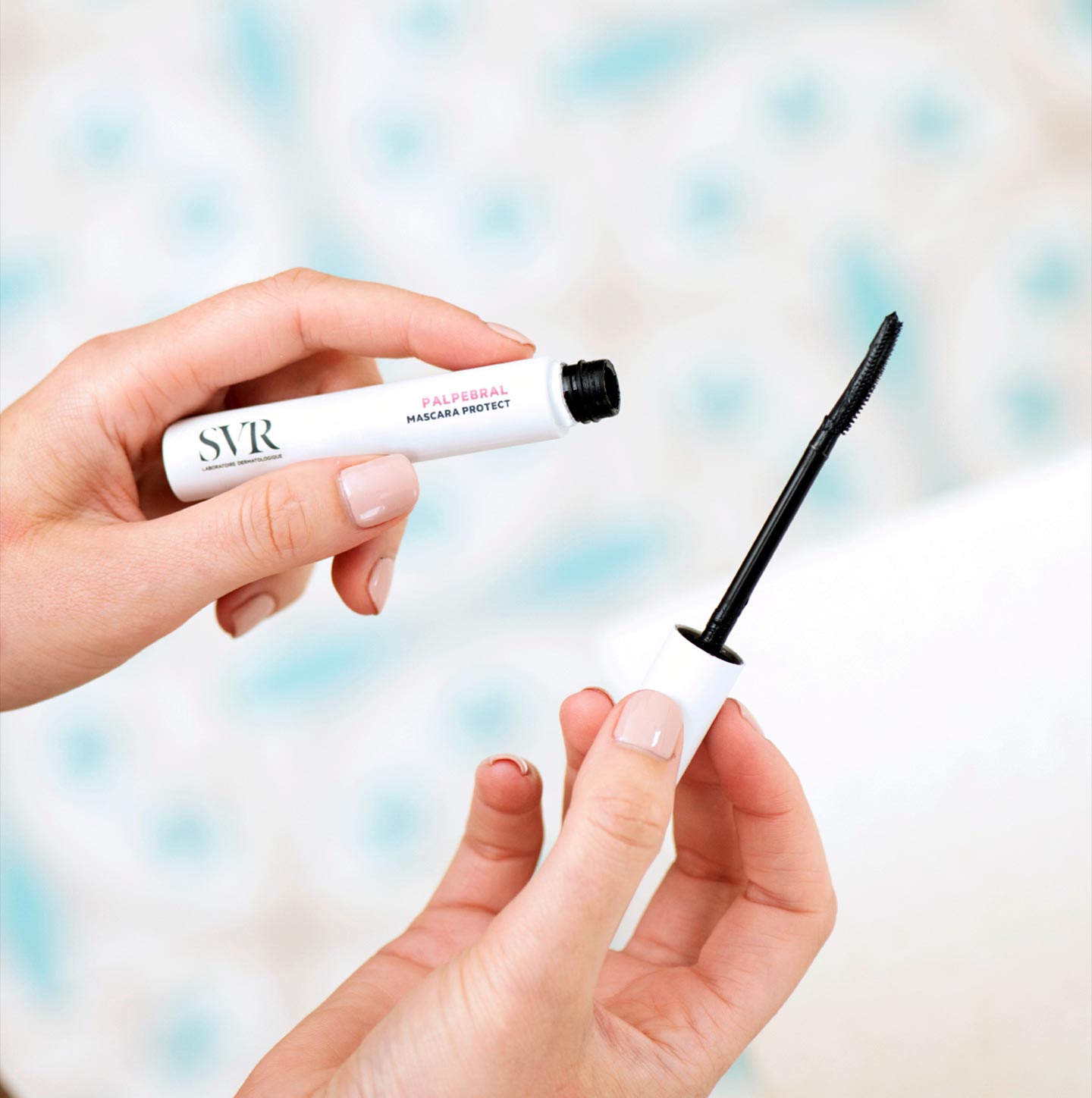 Hände halten die weiße SVR PALPEBRAL Mascara Protect Tube mit schwarzem Applikator vor einem hellen, gemusterten Hintergrund.
