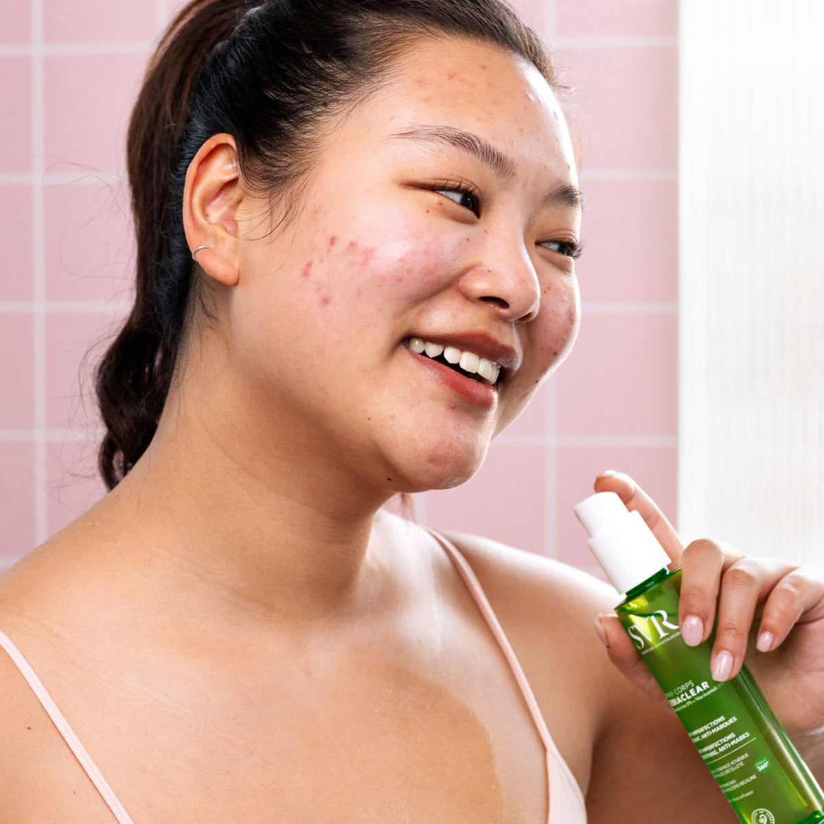 Eine junge Frau hält eine grüne SVR Sebiaclear Spray Corps Flasche vor einem rosa Kachelhintergrund.