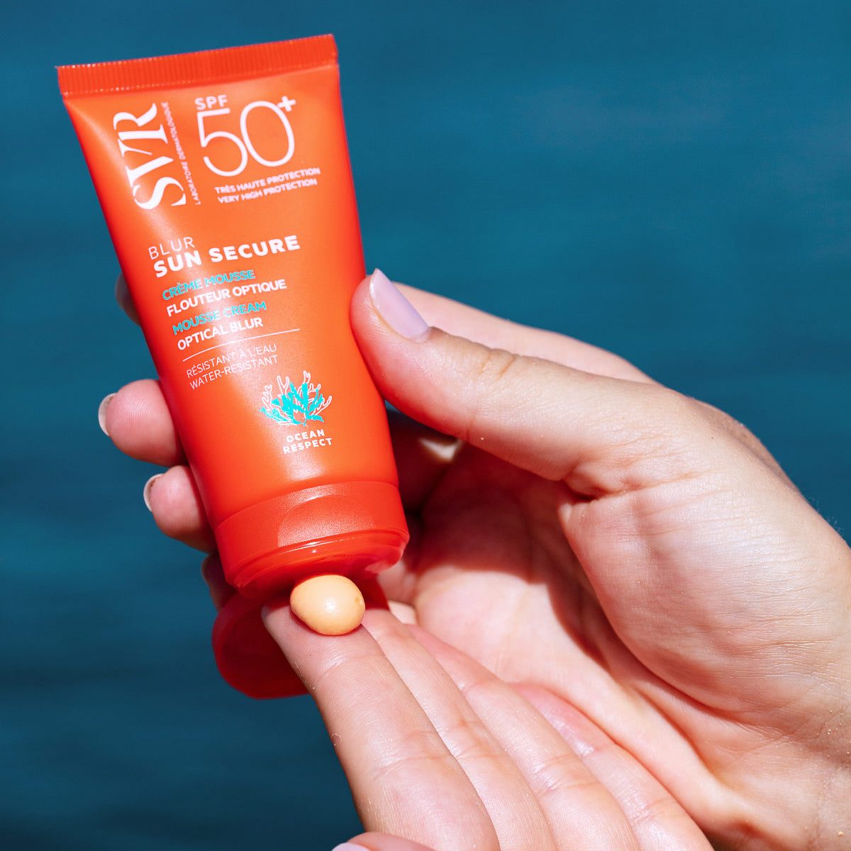 Orangefarbene SVR Sun Secure Blur SPF50+ Tube mit aprikosenfarbener Mousse-Creme auf einem Finger vor blauem Hintergrund.