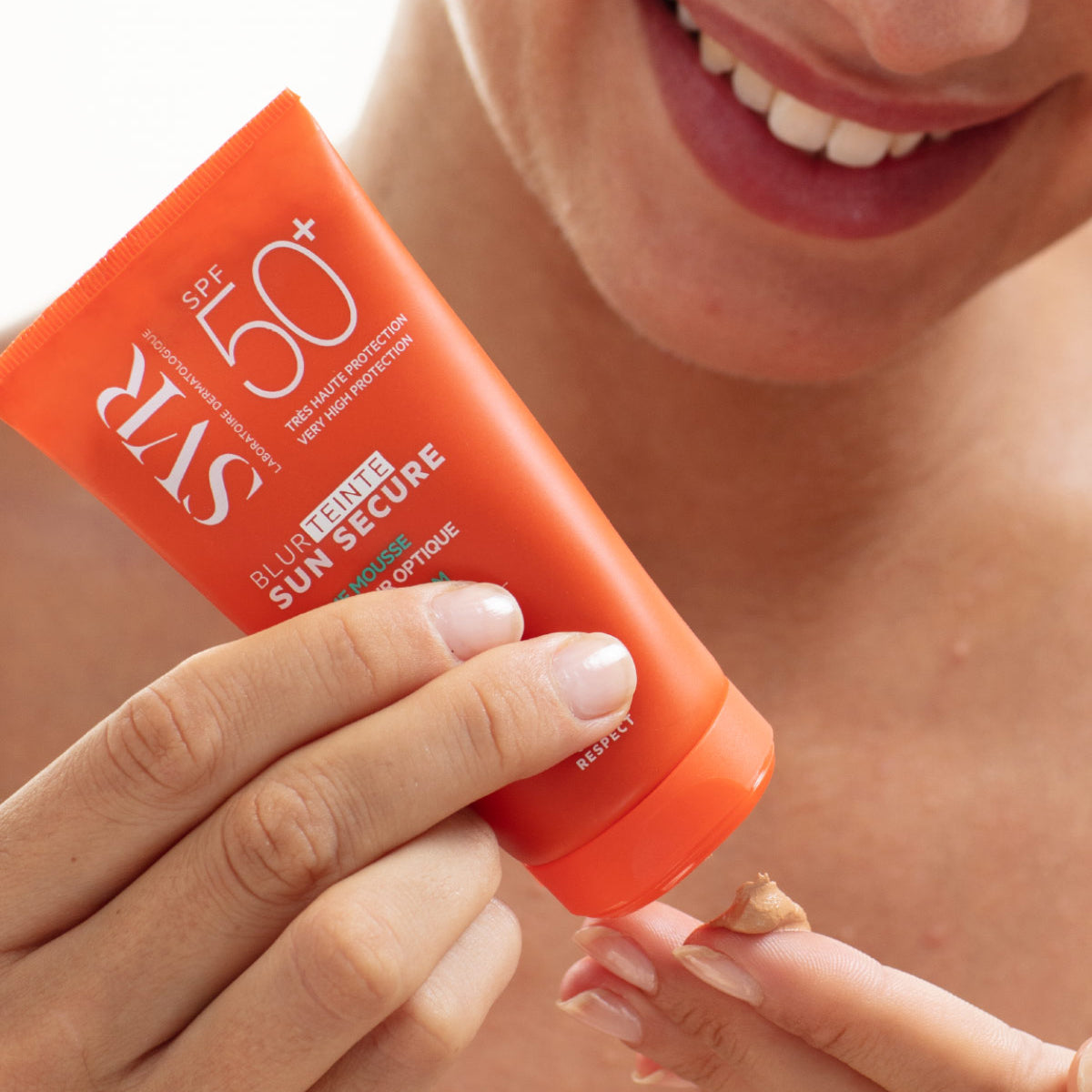 Orangefarbene SVR SUN SECURE Blur teinté SPF50+ Tube, aus der eine getönte Mousse-Creme auf den Finger gedrückt wird.