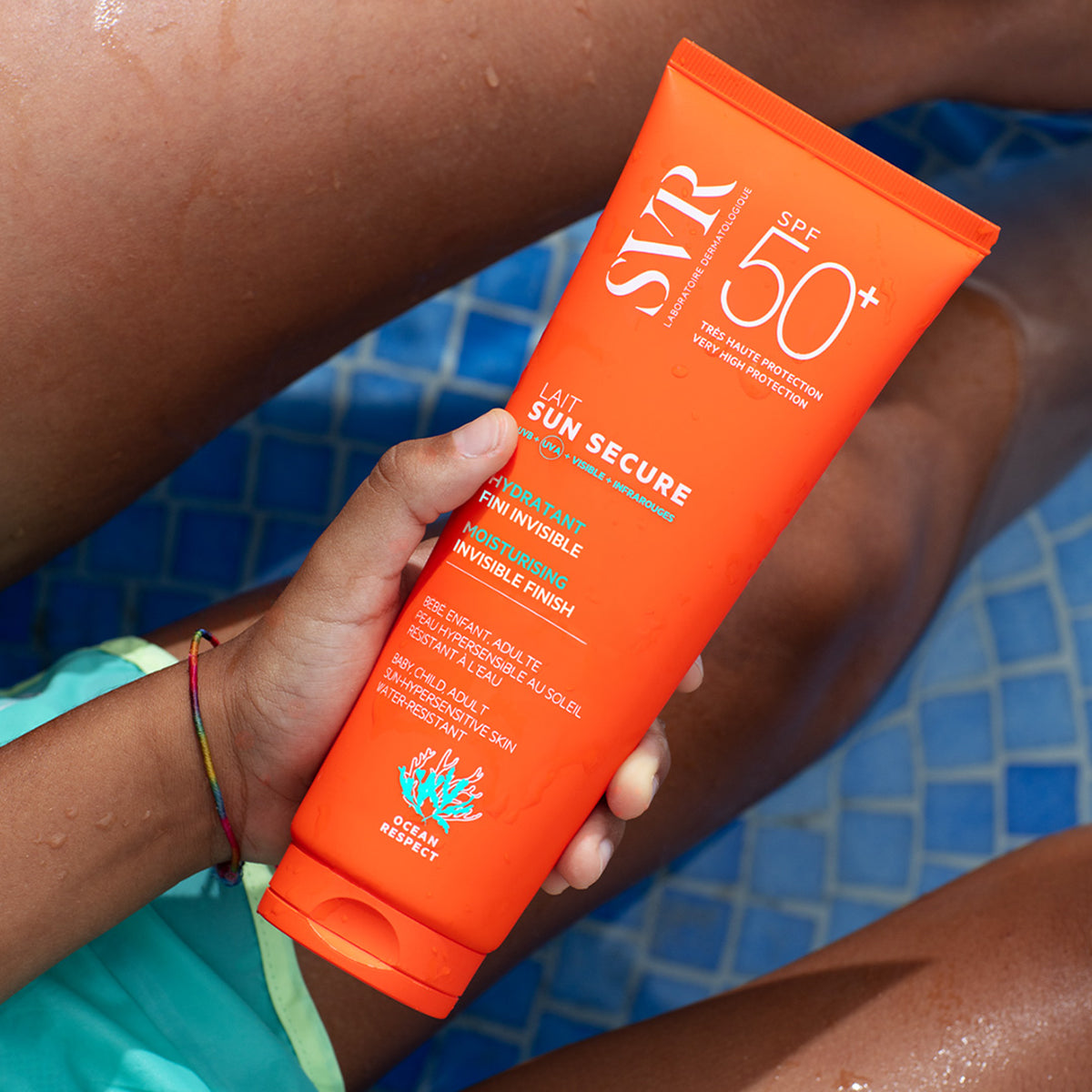 Hand hält orangefarbene SVR SUN SECURE Lait SPF50+ Sonnencreme-Tube vor einem blauen Poolhintergrund.