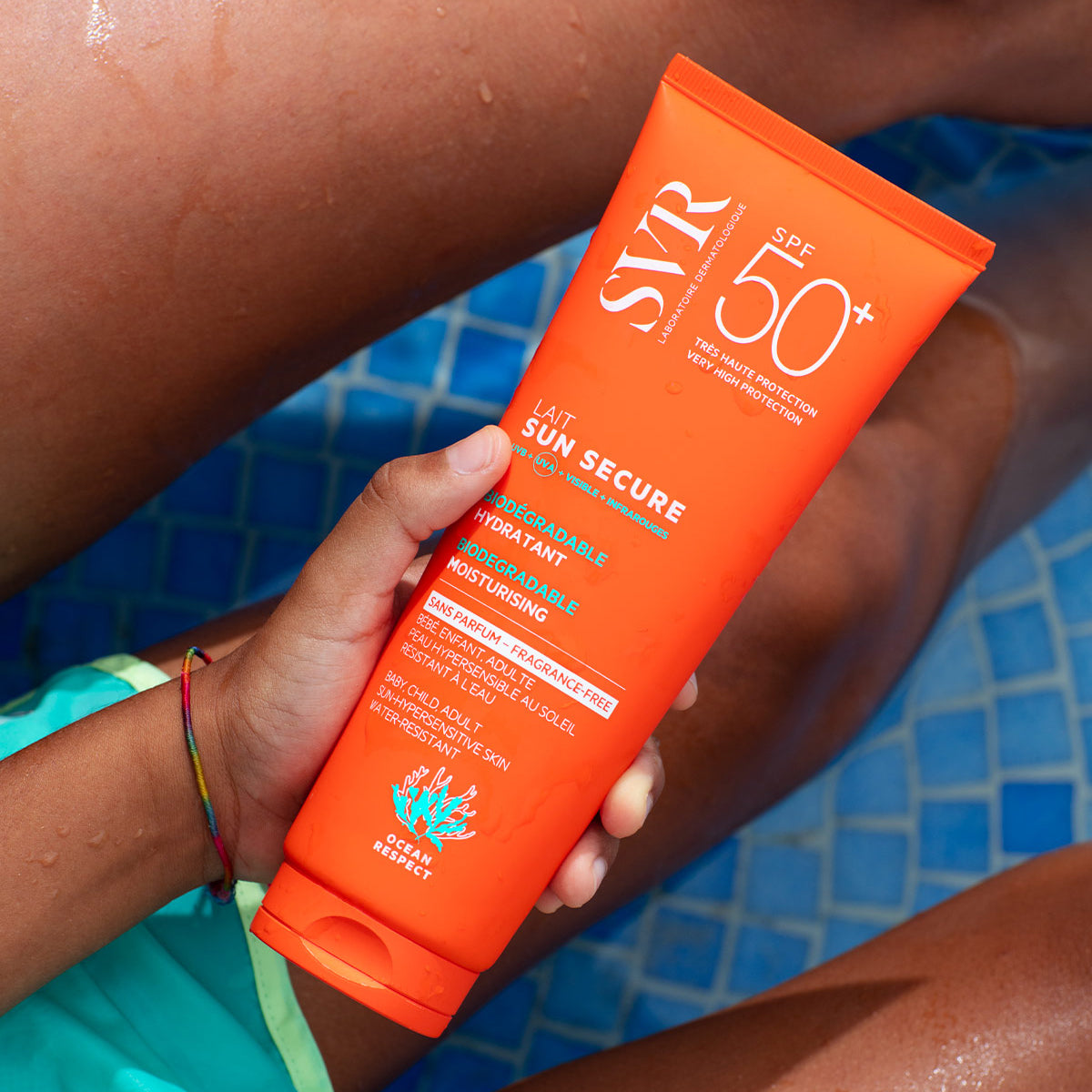 SVR SUN SECURE Lait SPF50+ Tube in leuchtendem Orange, gehalten von einer Hand vor einem blauen Poolhintergrund.