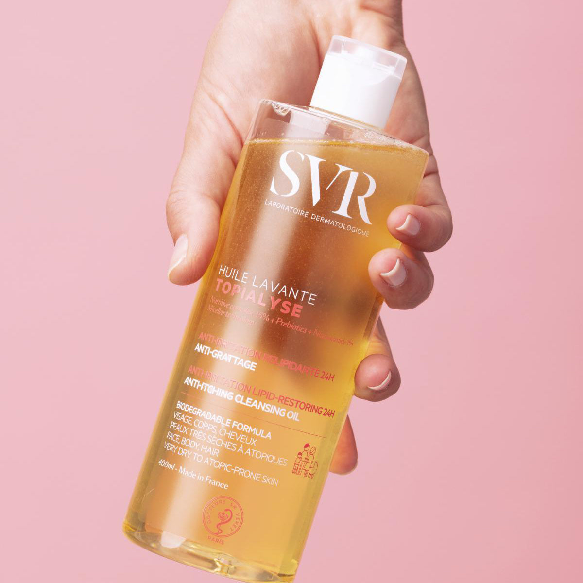 Hand hält eine transparente 400ml Flasche SVR TOPIALYSE Huile Lavante Duschöl mit weißem Verschluss vor rosa Hintergrund.