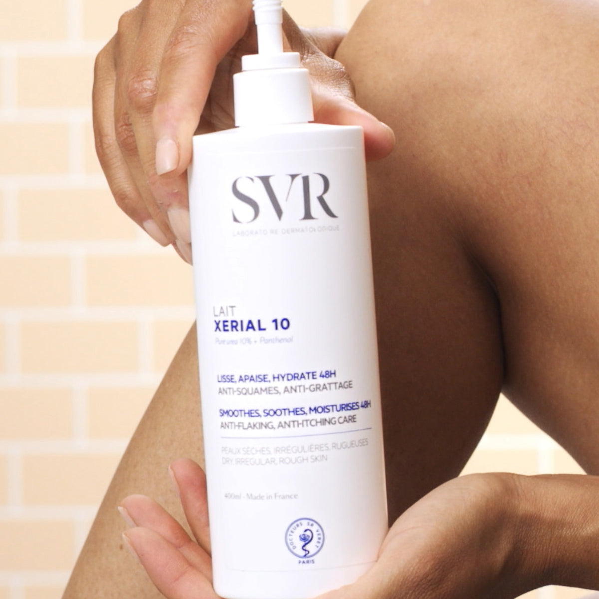 Weiße SVR XERIAL 10 Lait Pumpflasche mit blauer Beschriftung, gehalten vor einem Bein zur Pflege trockener, rauer Haut.