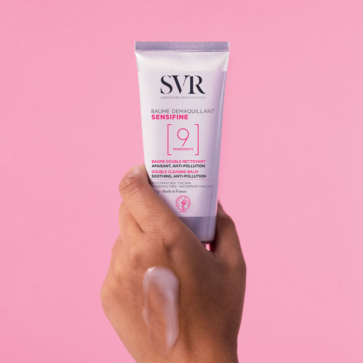 Hand hält weiße SVR Sensifine Reinigungsbalsam-Tube vor rosa Hintergrund mit einer öligen Texturprobe auf der Haut.