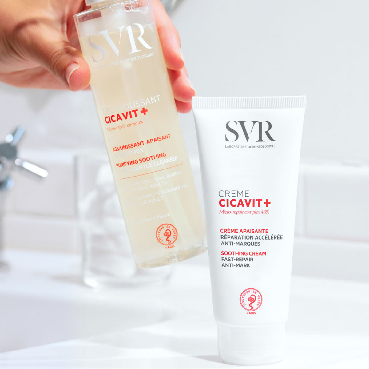 Hand hält SVR Cicavit+ Gel Moussant neben der Cicavit+ Creme in weißer Tube vor einem Badezimmerhintergrund.