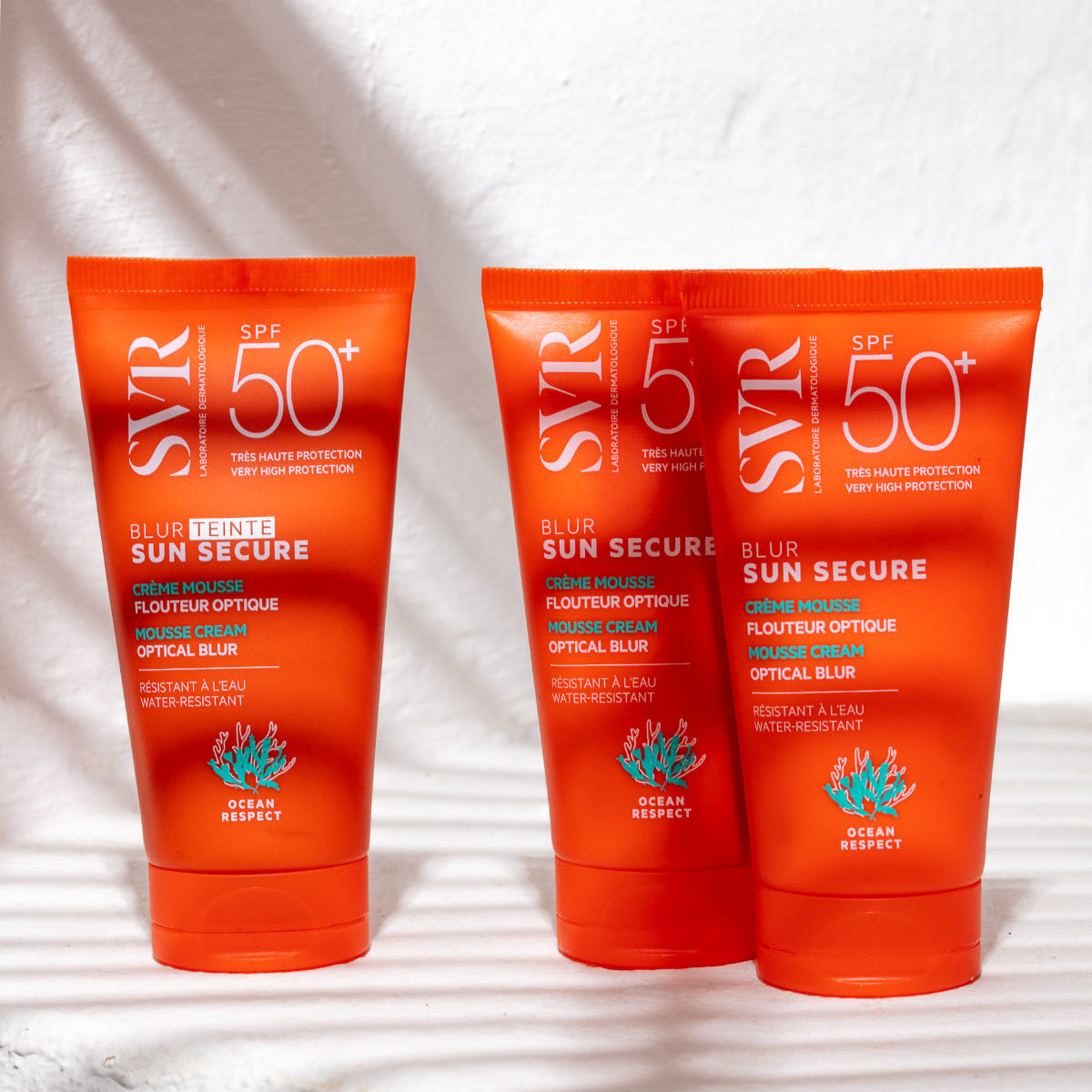 Drei orangefarbene SVR SUN SECURE Blur SPF50+ Sonnencreme-Tuben mit weißer Schrift auf weißem Untergrund mit Schattenwurf.