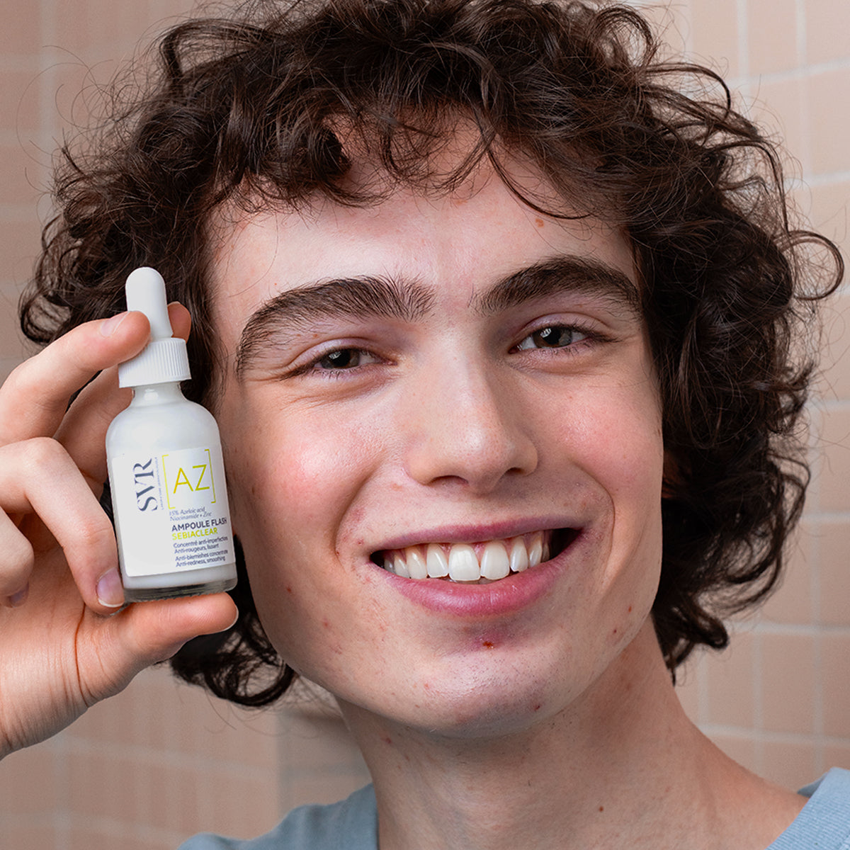 Lächelnder junger Mann hält eine weiße SVR Sebiaclear Ampoule Flash Glasflasche mit Pipette vor die Kamera.