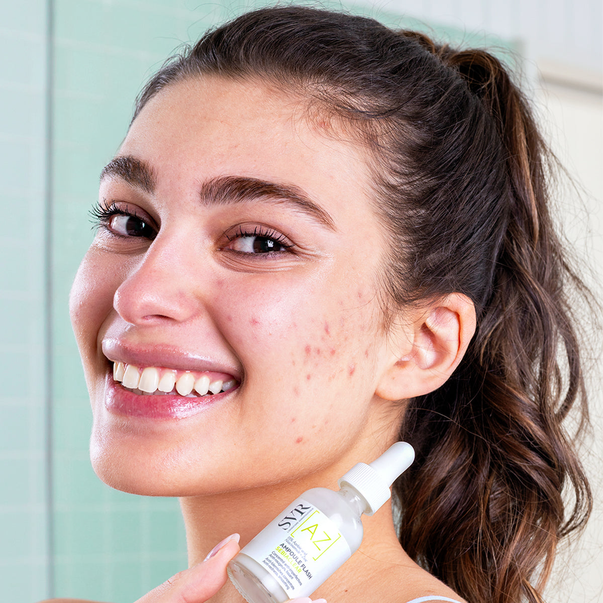 Smiling woman holding an SVR Ampoule Flash serum bottle for acne treatment against a light green tiled background.