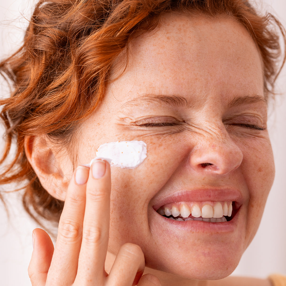 Woman with red hair and freckles applying white, textured C20 Biotic cream to her cheek with a smile.