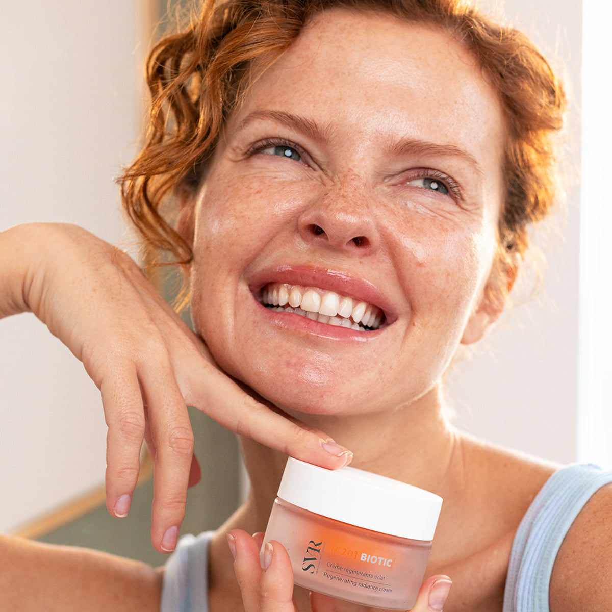 Smiling woman with freckles holding a frosted glass jar of SVR C20 Biotic regenerating radiance cream.