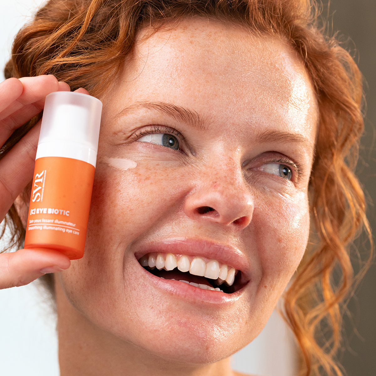 Woman holding an orange SVR C-EYE Biotic pump bottle with eye cream applied to her cheek.