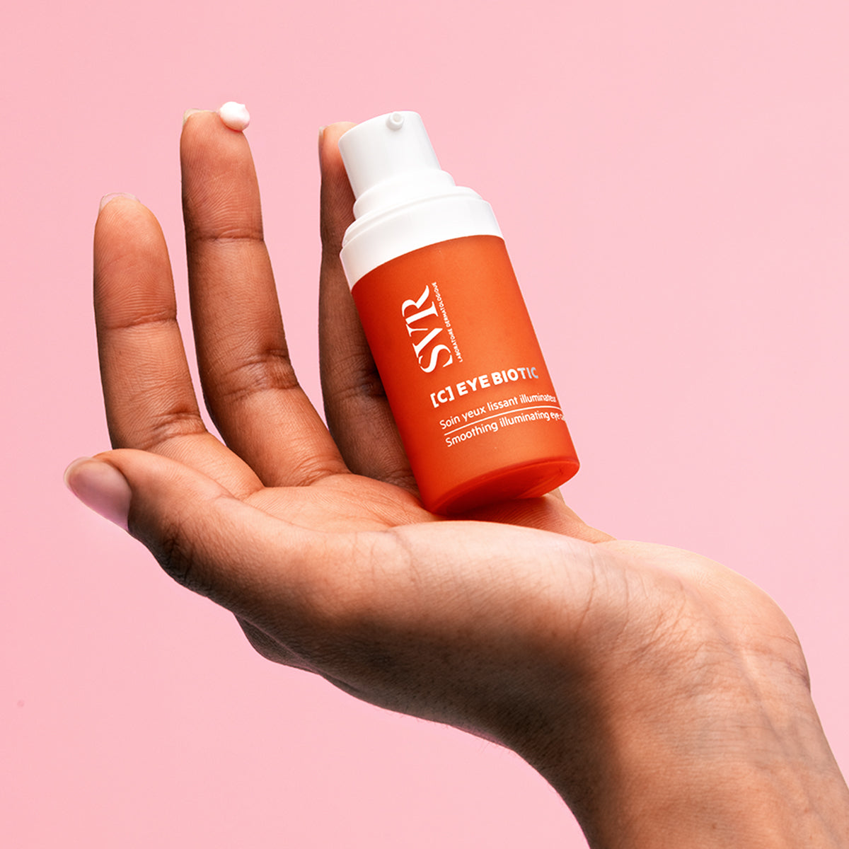 SVR C-EYE Biotic smoothing eye cream in an orange pump bottle held in a hand with a dab of white cream on a fingertip.