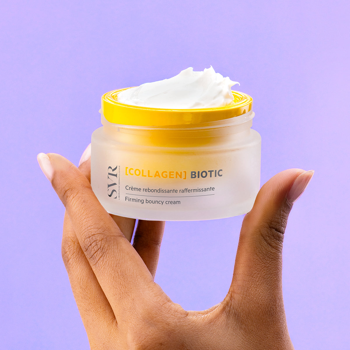 Hand holding a frosted glass jar of SVR Collagen Biotic firming cream with a bright yellow lid and white textured cream.