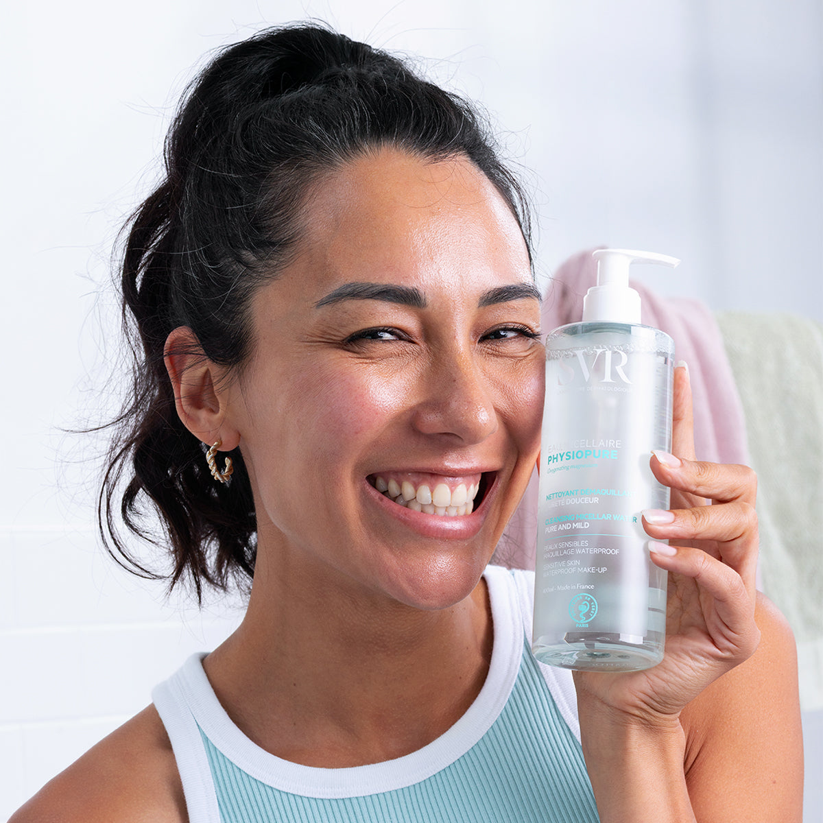 Lächelnde Frau hält eine transparente SVR PHYSIOPURE Mizellenwasser-Flasche mit weißem Pumpspender in die Kamera.