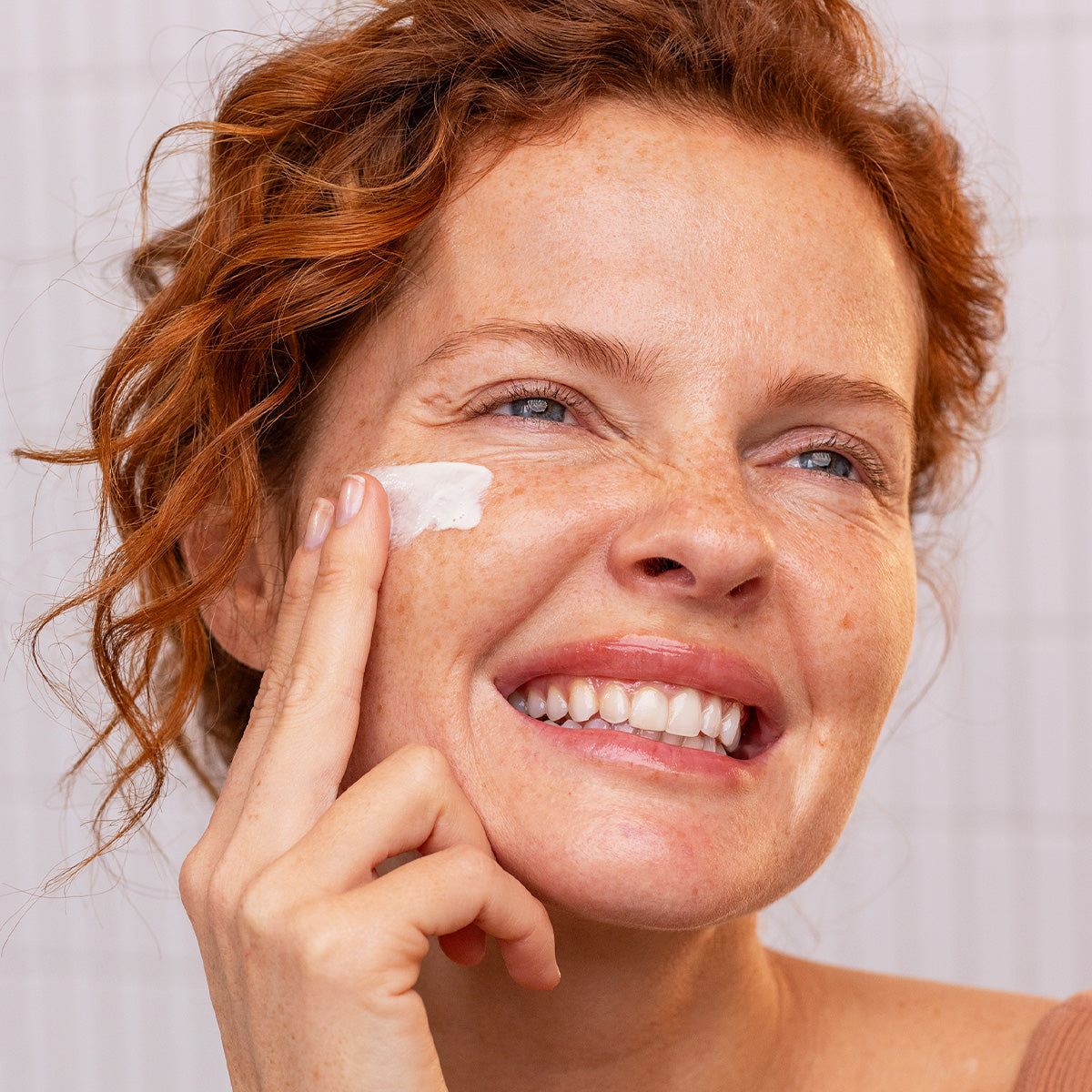Frau mit rötlichem Haar trägt das SUN SECURE Fluide SPF50+ auf ihre Wange auf und lächelt strahlend in die Kamera.