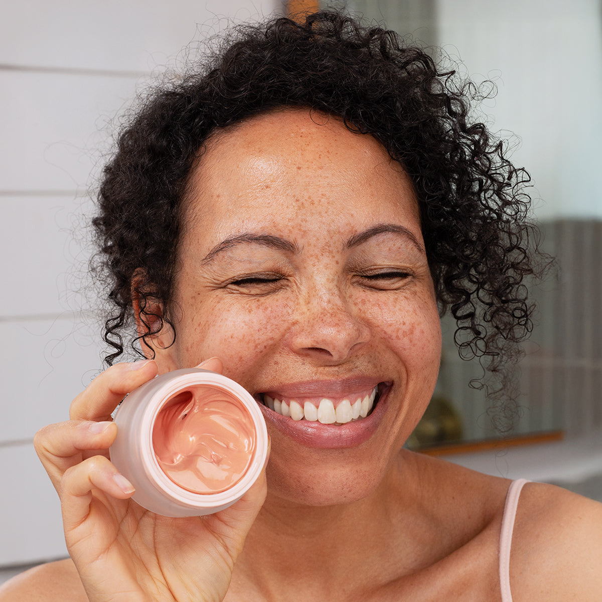 Smiling woman holding a jar of Densitium Rose Éclat cream with a smooth, pink-tinted texture in a frosted glass jar.