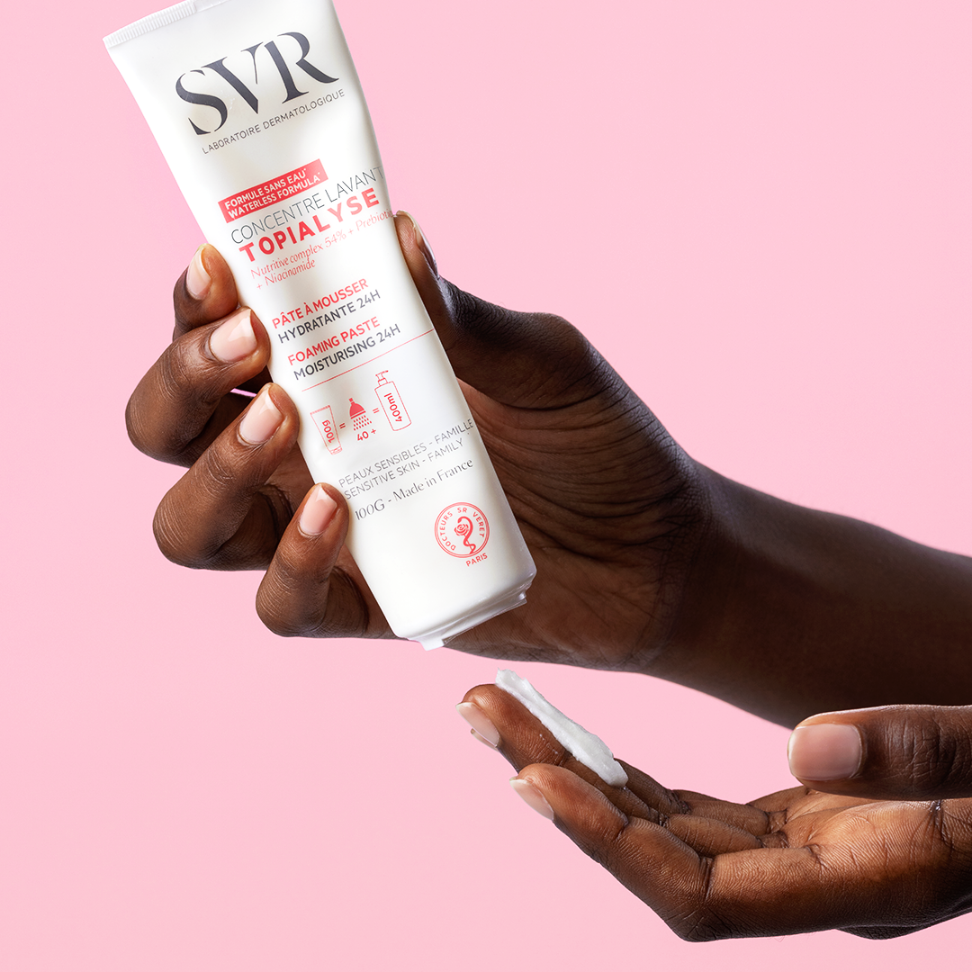 Hand holding a white 100g tube of SVR Topialyse Concentré lavant, dispensing a small amount of white foaming paste.