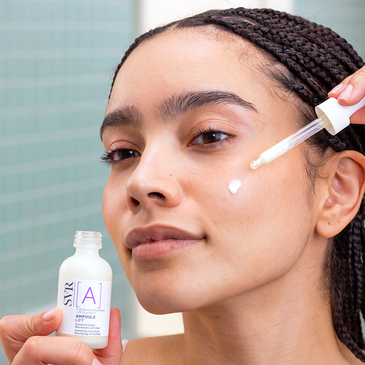 Woman applying SVR Ampoule Lift serum with a glass dropper to her cheek from a small white bottle.