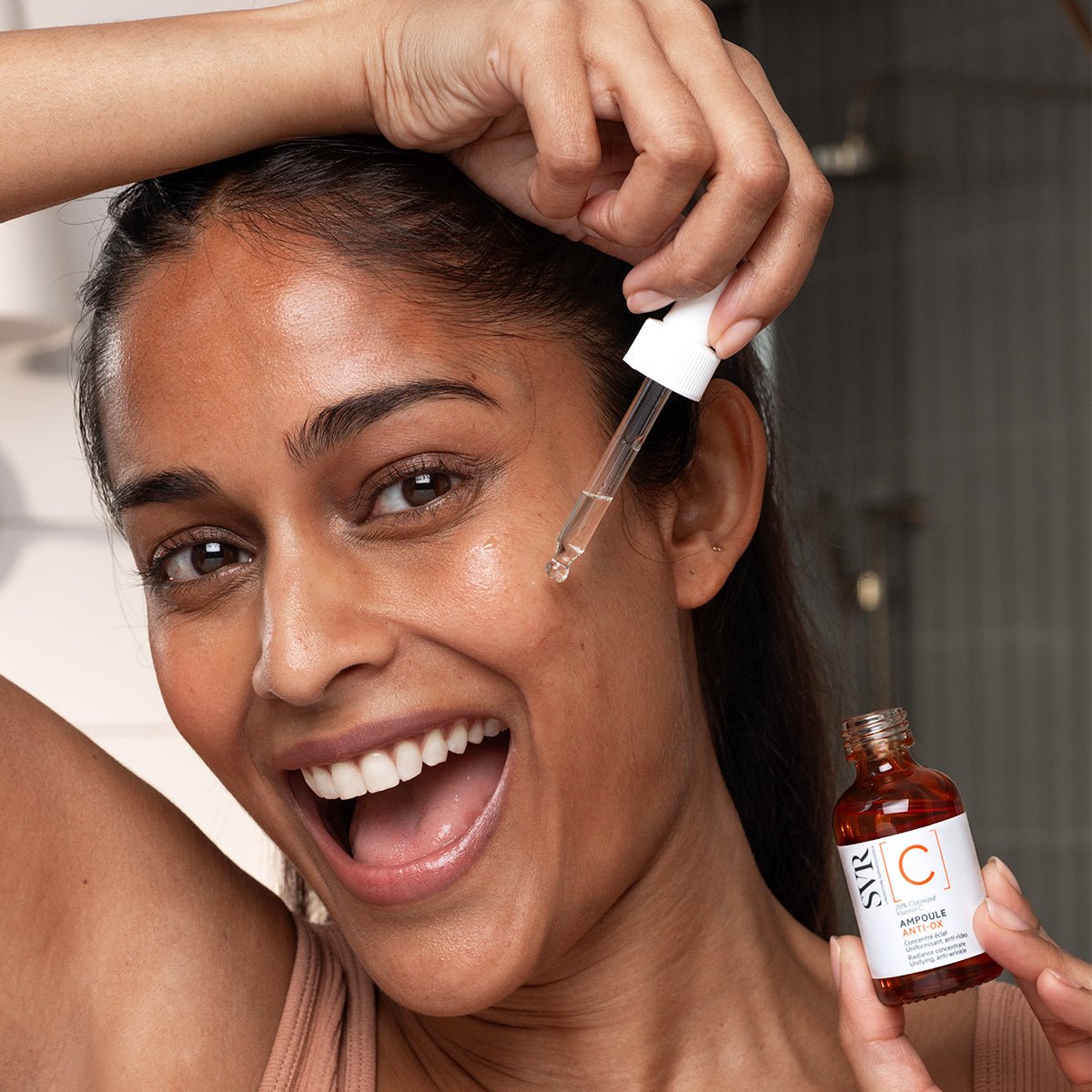 Lächelnde Frau trägt SVR Ampoule Anti-Ox Vitamin C Serum mit Pipette auf ihr Gesicht auf, braune Glasflasche in der Hand.