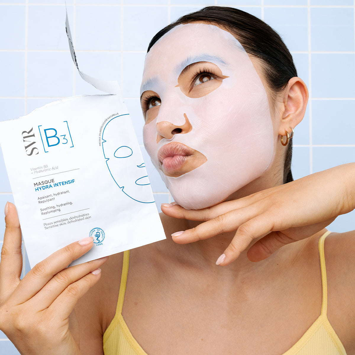 Frau mit weiĂer Biozellulose-Gesichtsmaske hält SVR B3 Masque Hydra Intensif Verpackung vor hellblauem Fliesen-Hintergrund.