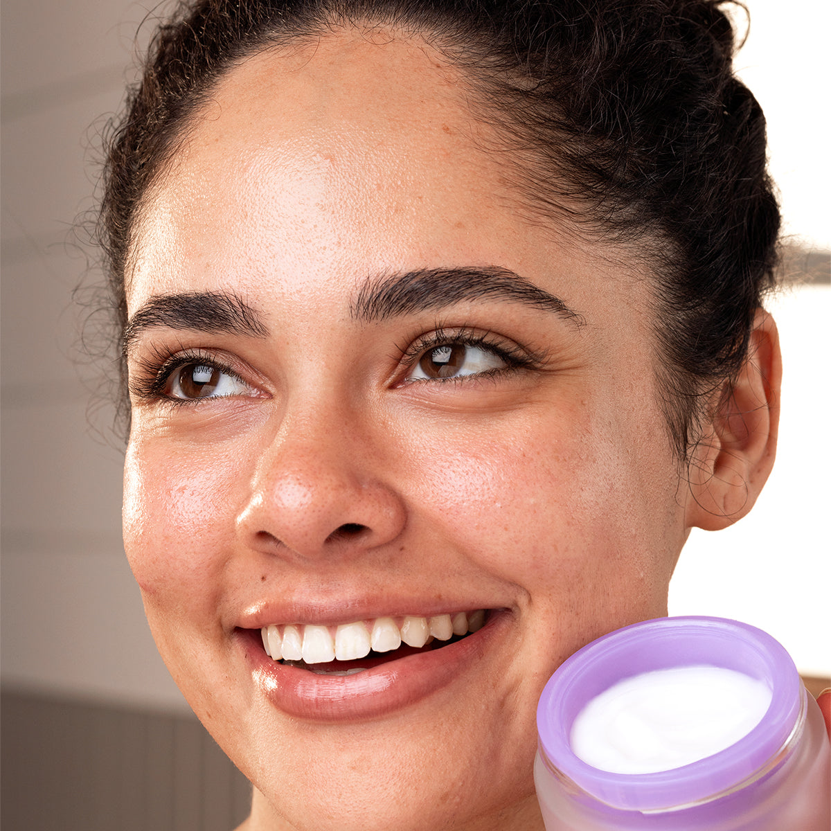 Smiling woman holding a frosted glass jar of PEPTI Biotic cream with a purple lid and white moisturizer.