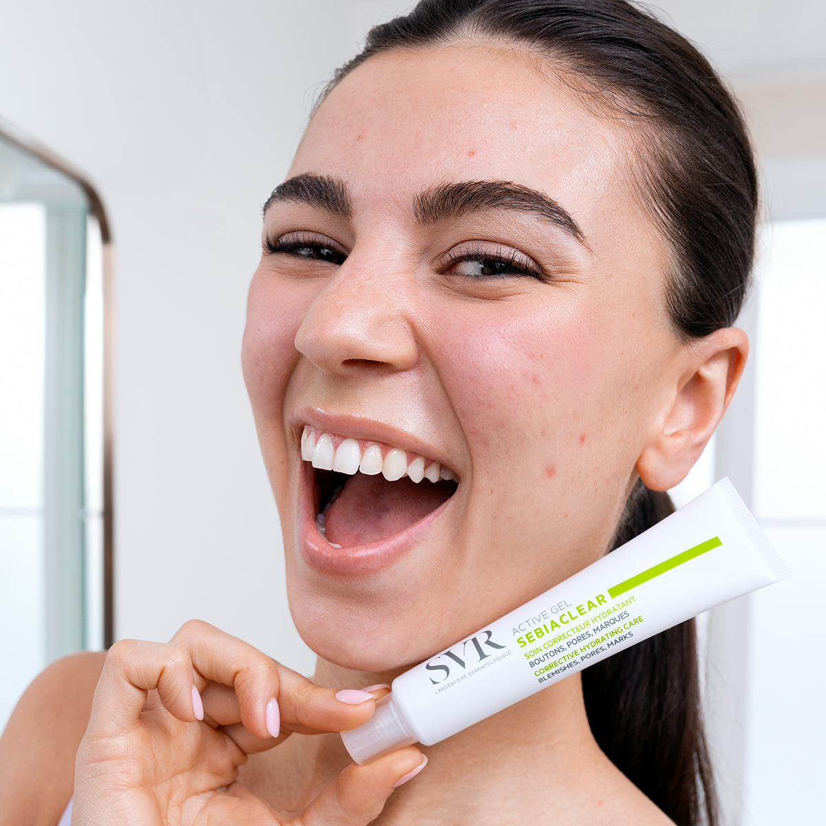 Smiling woman holding a white SVR Sebiaclear Active Gel tube with green accents in a bright bathroom setting.