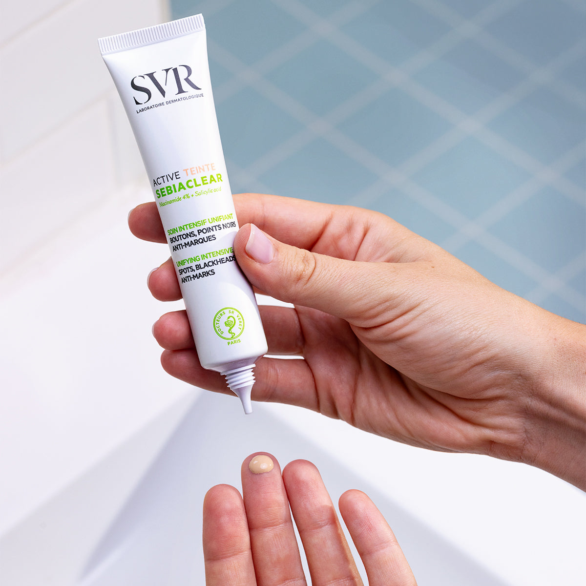 Weiße SVR Sebiaclear Active Teinté Tube in der Hand, ein Klecks der getönten Creme auf dem Finger vor einem Badezimmerhintergrund.
