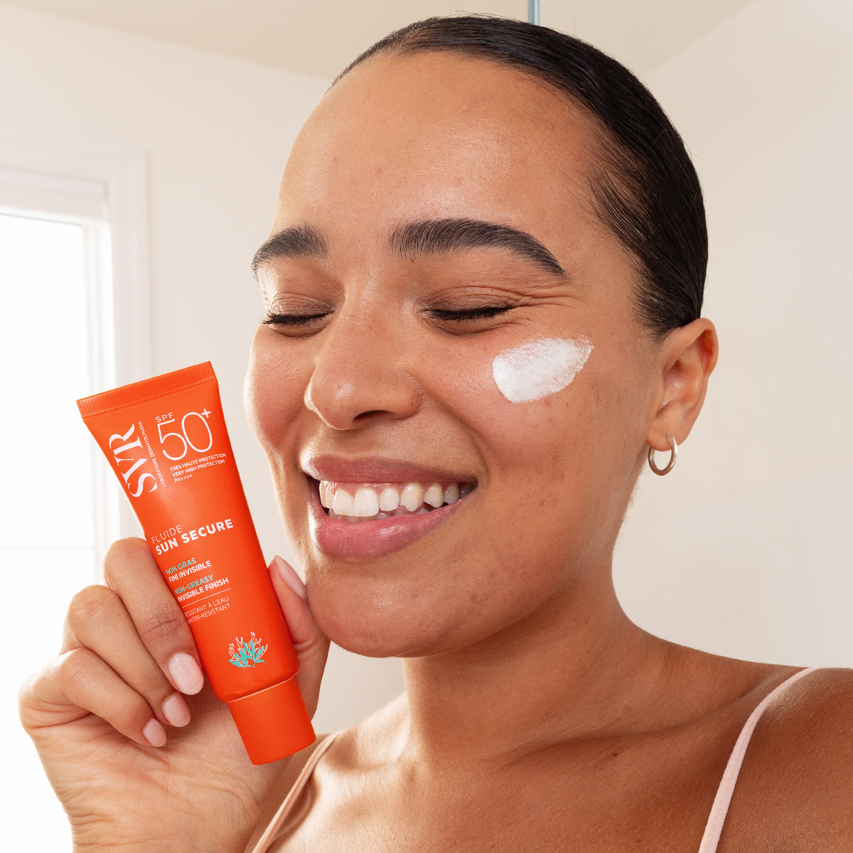 Frau hält eine orangefarbene SVR SUN SECURE Fluide SPF50+ Tube mit weißer Sonnencreme auf der Wange.