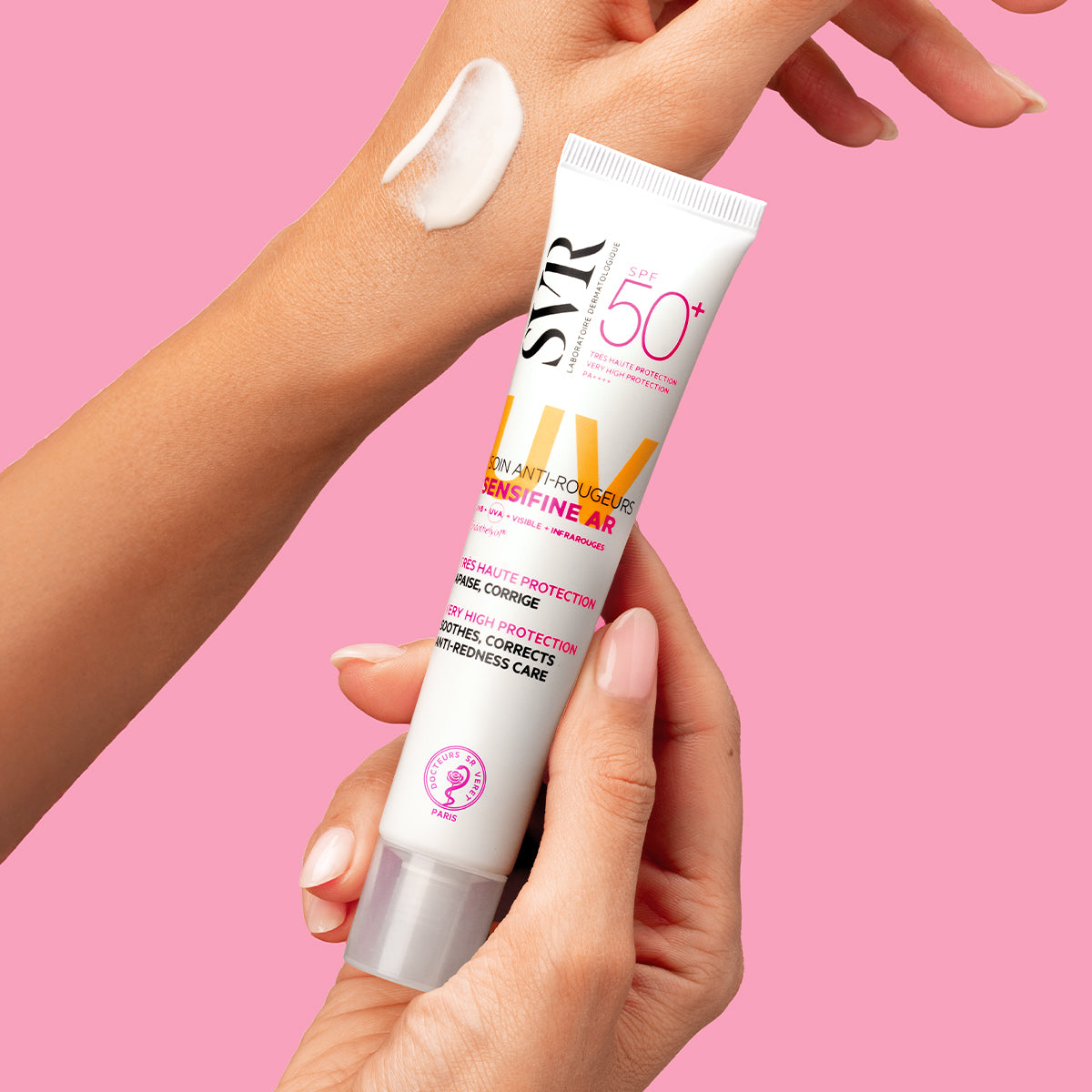 SVR Sensifine AR Crème SPF50+ Tube in Händen gehalten, mit weißer Textur auf dem Handrücken vor rosa Hintergrund.