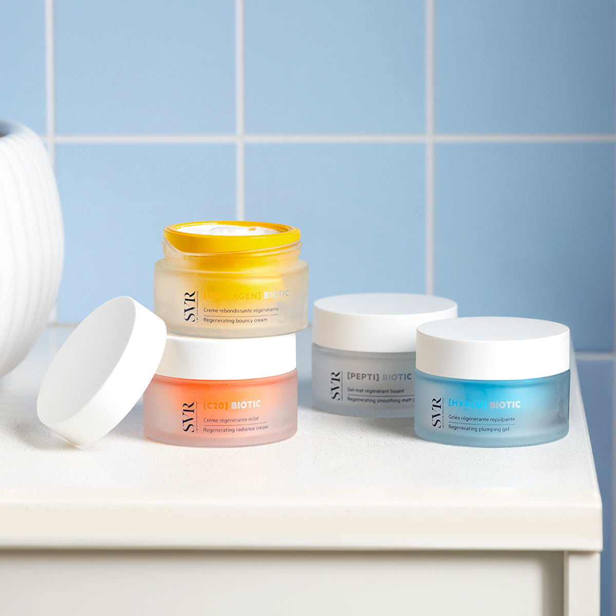 SVR Biotic skincare jars in yellow, orange, grey, and blue on a white bathroom counter against blue tiles.