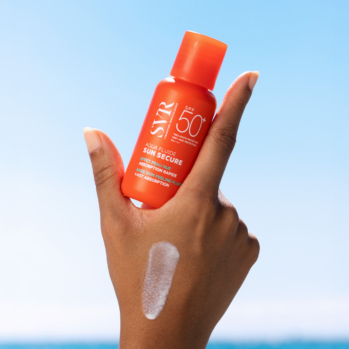 Hand holding an orange bottle of SVR SUN SECURE Aqua Fluide SPF50+ with a streak of white sunscreen on the skin.