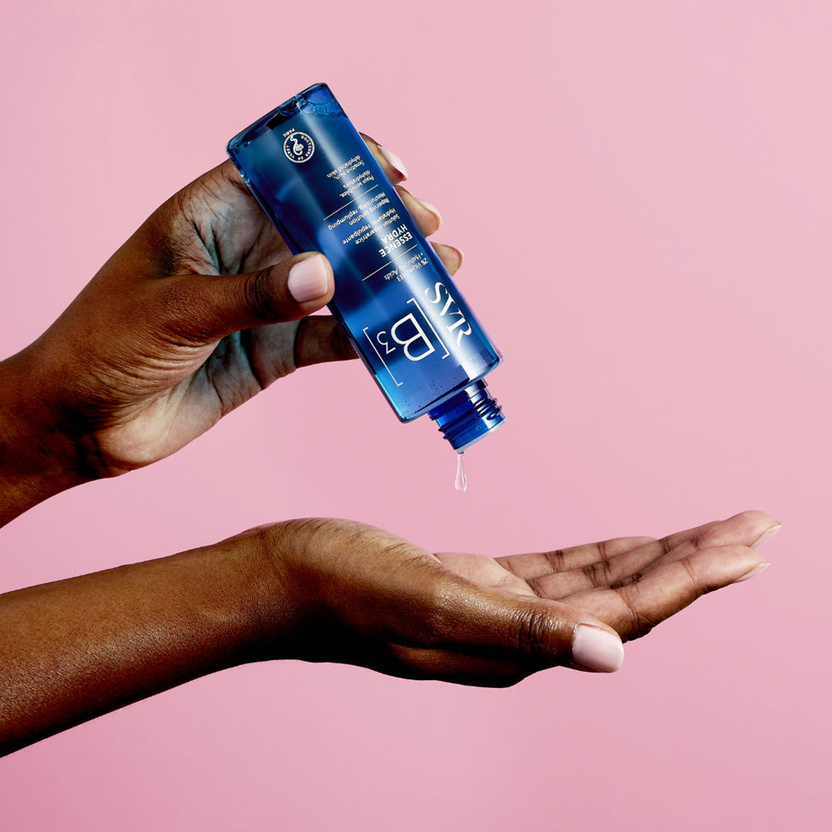 Blaue SVR B3 Essence Hydra Flasche, die eine transparente Flüssigkeit in eine dunkle Handfläche tropft, vor rosa Hintergrund.