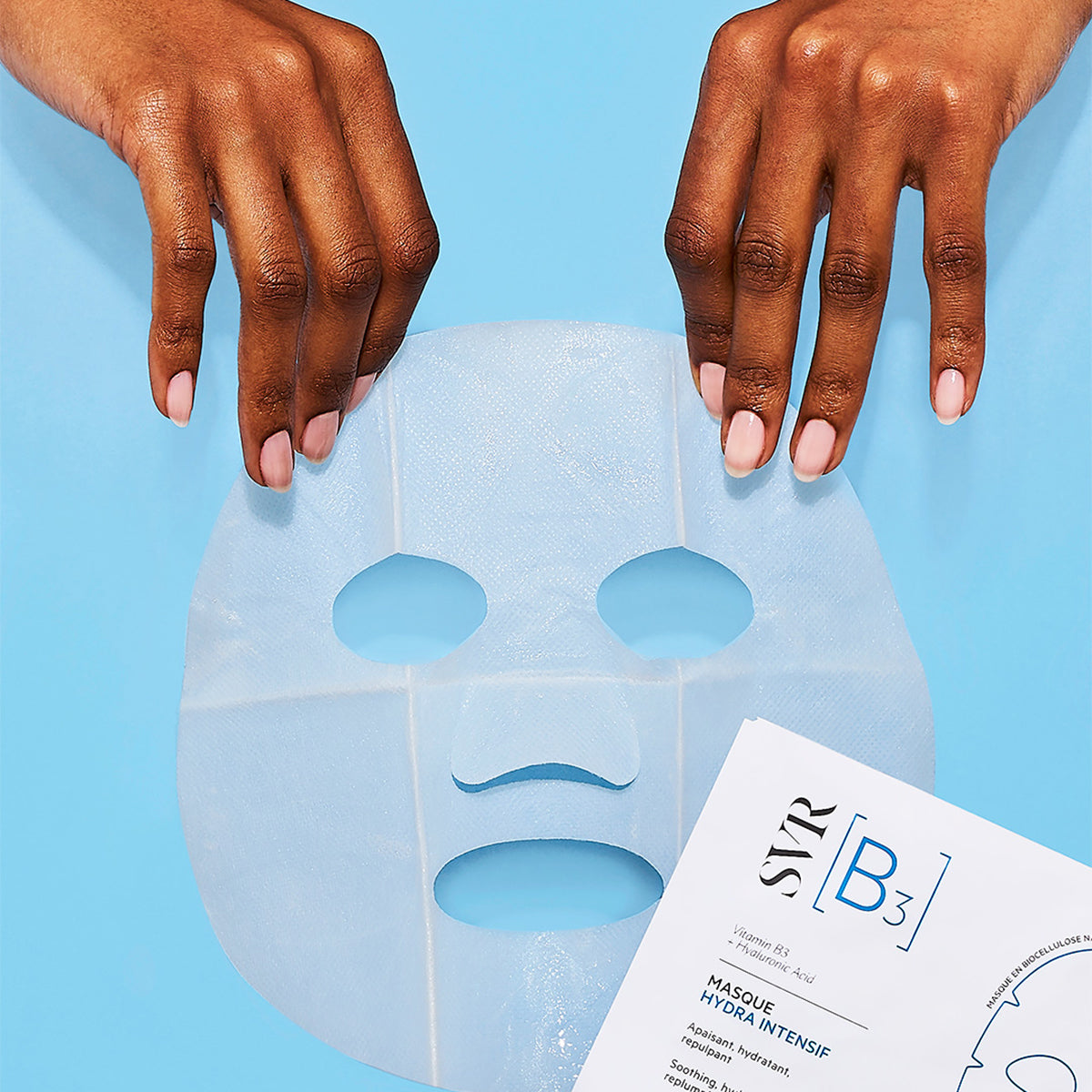 Hände halten eine feuchte, halbtransparente Biozellulose-Gesichtsmaske vor hellblauem Hintergrund neben der SVR B3 Maskenverpackung.