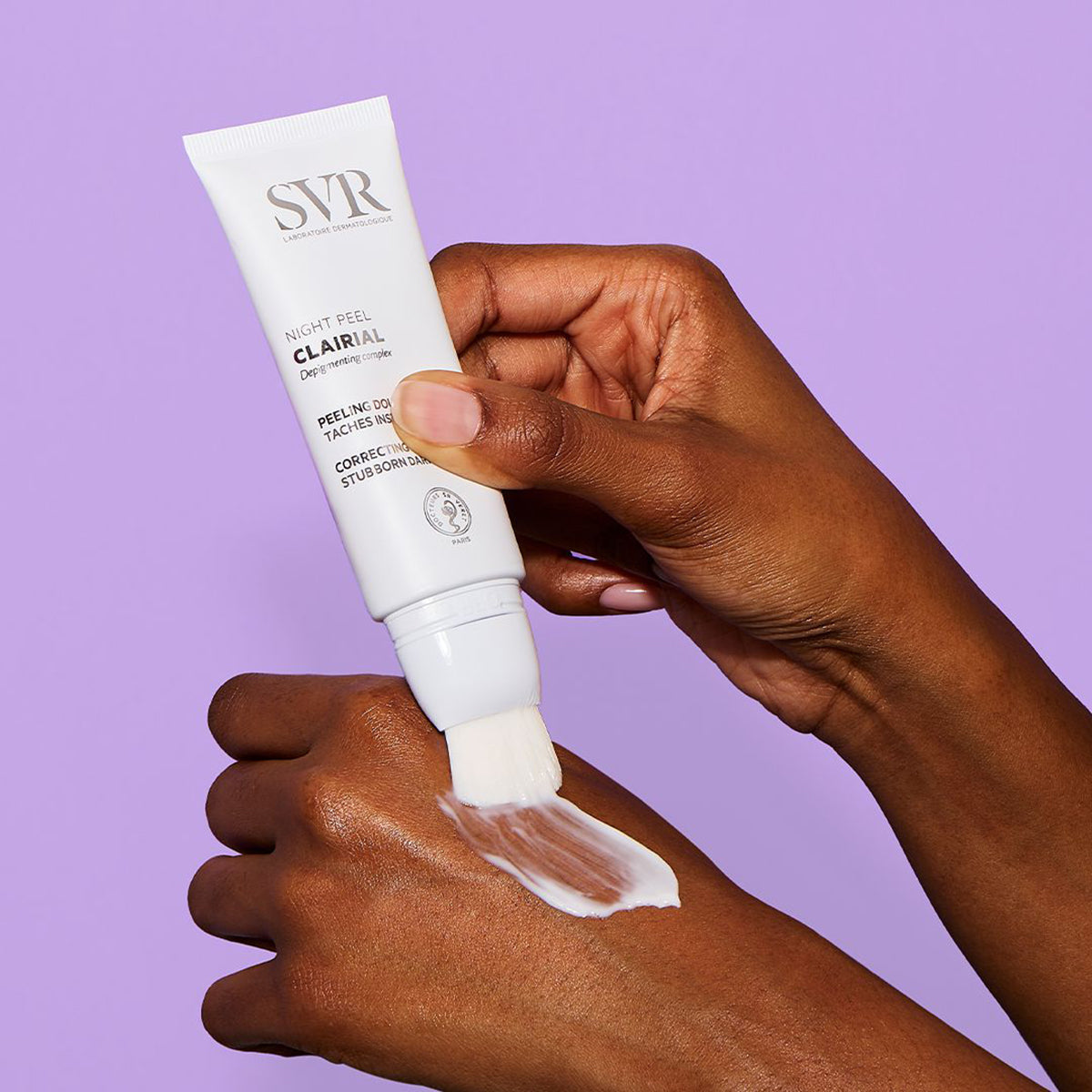 SVR Clairial Night Peel Tube mit Pinselapplikator beim Auftragen einer weißen Peeling-Creme auf den Handrücken.