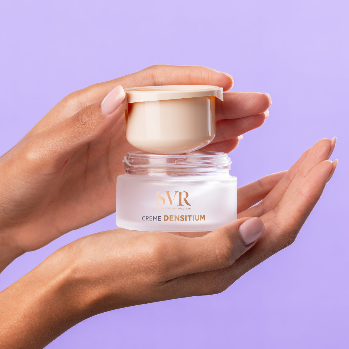 Hands holding an open jar of SVR DENSITIUM Crème with a frosted glass base and beige lid against a purple background.