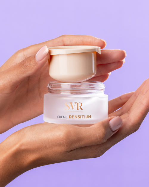 Hands holding an open jar of SVR DENSITIUM Crème with a frosted glass base and beige lid against a purple background.