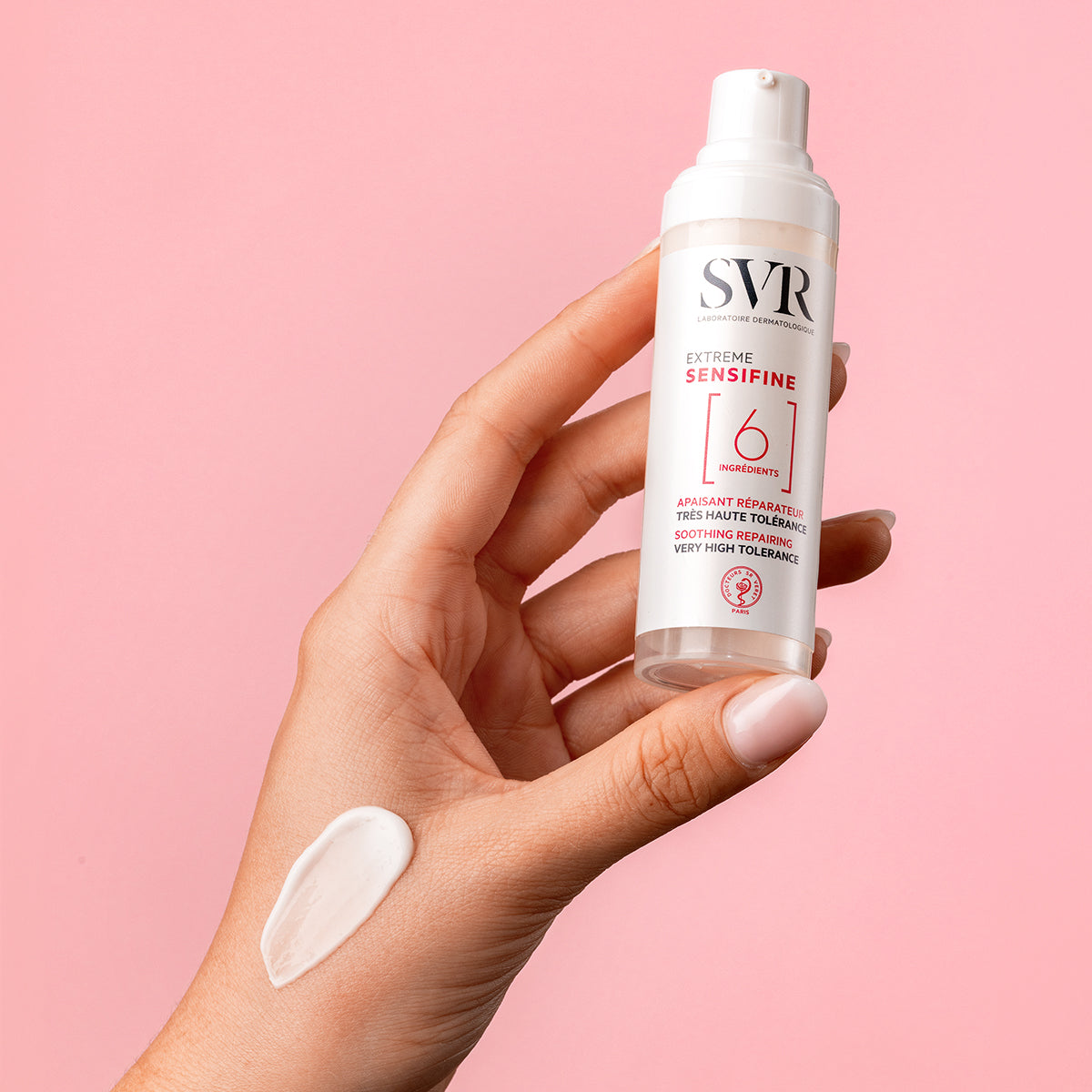 Hand holding a white SVR Sensifine Extreme pump bottle with a dab of cream on the skin against a pink background.