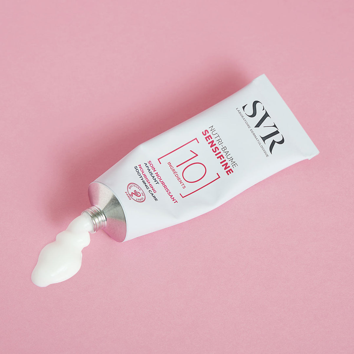 Weiße SVR SENSIFINE Nutri-Baume Aluminiumtube mit austretender cremiger Textur auf einem rosa Hintergrund.